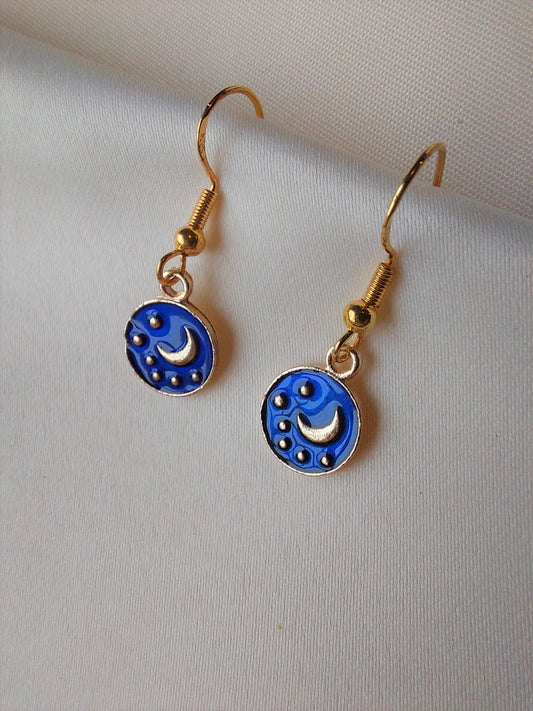 Small blue and gold earrings. Circle earring with moon and stars inside. Gold hooks. Earrings laid out on fabric.