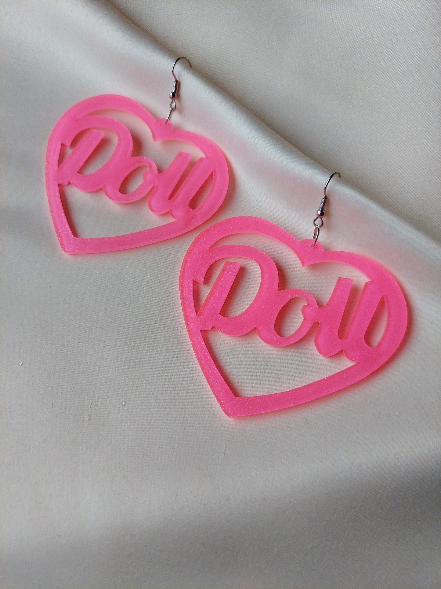 Bright pink heart shaped earrings with doll lettering in the middle of the heart. Silver earring hooks in the middle of the heart. Pink include some glitter throughout it.