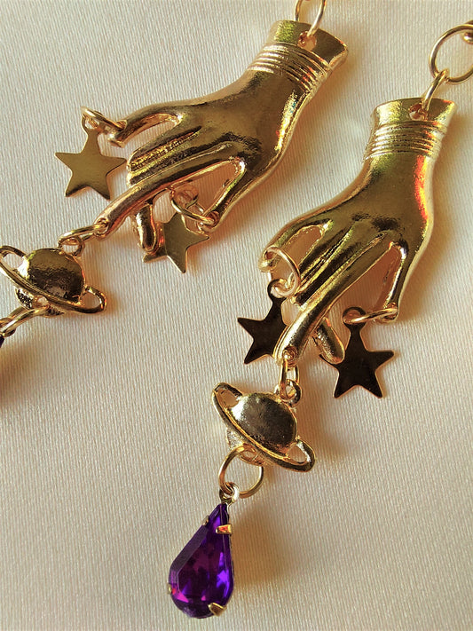 Gold earrings. Hand charm pointing down, gold stars on finger tips followed by Saturn planet charm. Underneath plant charm, there is a purple tear drop shaped gem. Earrings are all gold, including hooks except for purple gem.