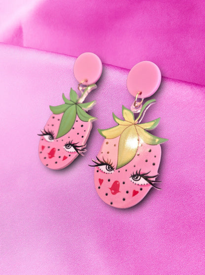Strawberry-shaped earrings that have a face on them with pink and green details on a pink background