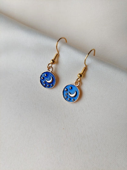 Small blue and gold earrings. Circle earring with moon and stars inside. Gold hooks. Earrings laid out on fabric. 