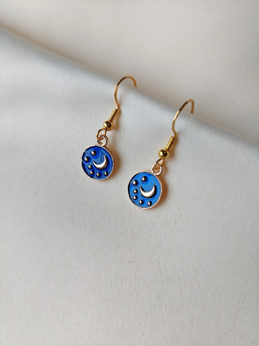 Small blue and gold earrings. Circle earring with moon and stars inside. Gold hooks. Earrings laid out on fabric. 