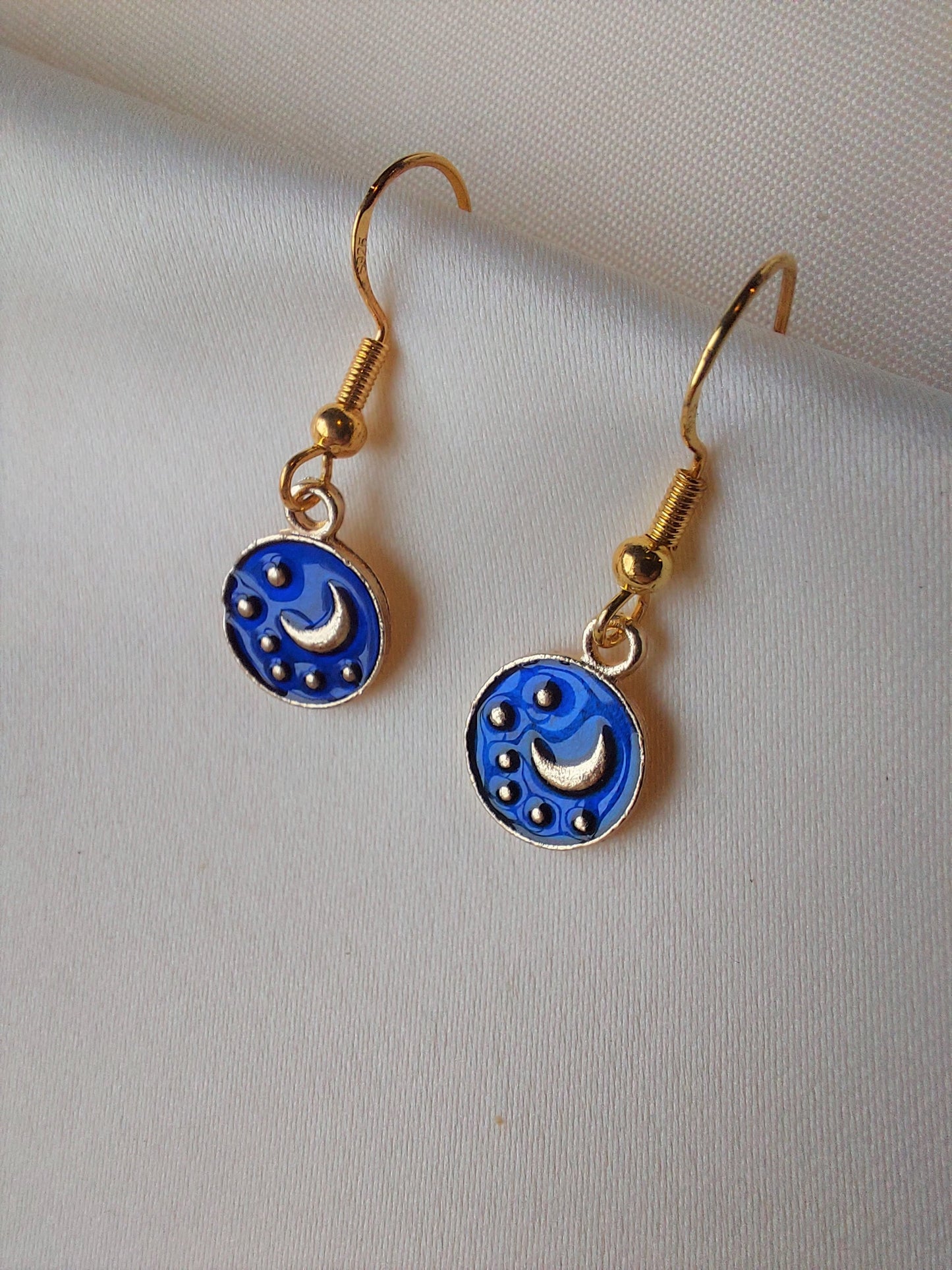 Small blue and gold earrings. Circle earring with moon and stars inside. Gold hooks. Earrings laid out on fabric. 