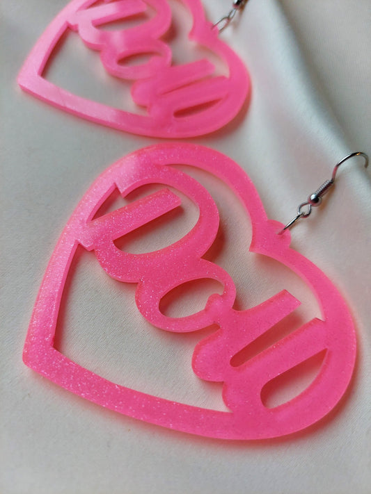 Bright pink heart shaped earrings with doll lettering in the middle of the heart. Silver earring hooks in the middle of the heart. Pink include some glitter throughout it. 