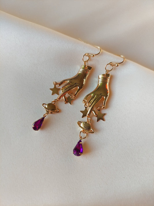 Gold earrings.  Hand charm pointing down, gold stars on finger tips followed by Saturn planet charm. Underneath plant charm, there is a purple tear drop shaped gem. Earrings are all gold, including hooks except for purple gem. These earrings are dangly but not too long. 