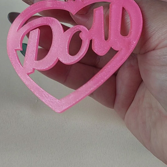 Bright pink heart shaped earrings with doll lettering in the middle of the heart. Silver earring hooks in the middle of the heart. Pink include some glitter throughout it. Held in hand for size comparison. 