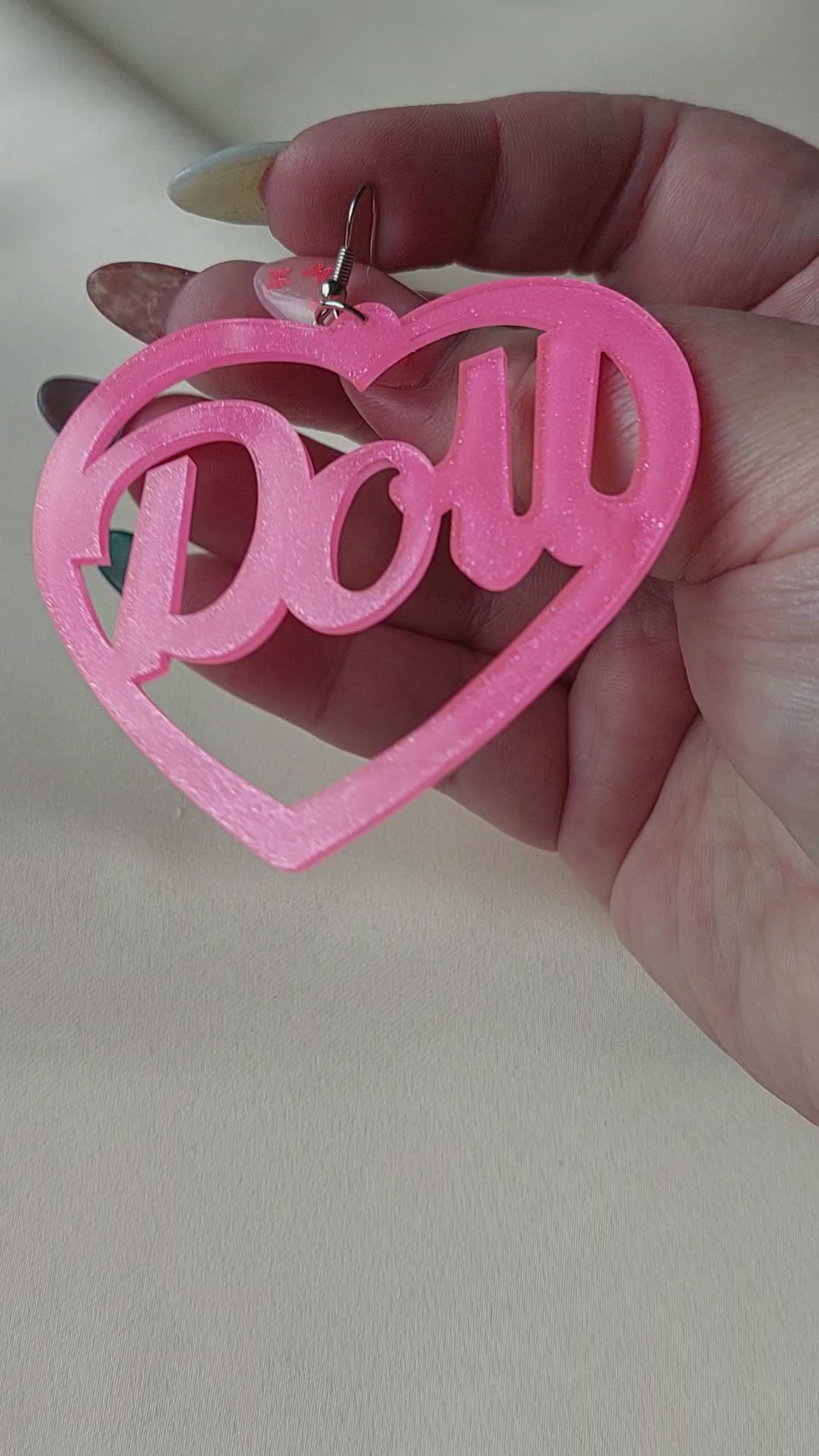 Bright pink heart shaped earrings with doll lettering in the middle of the heart. Silver earring hooks in the middle of the heart. Pink include some glitter throughout it. Held in hand for size comparison. 