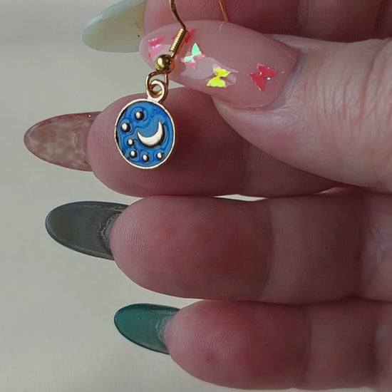 Small blue and gold earrings. Circle earring with moon and stars inside. Gold hooks. Earrings laid out on fabric. Hand holding for size comparison. 