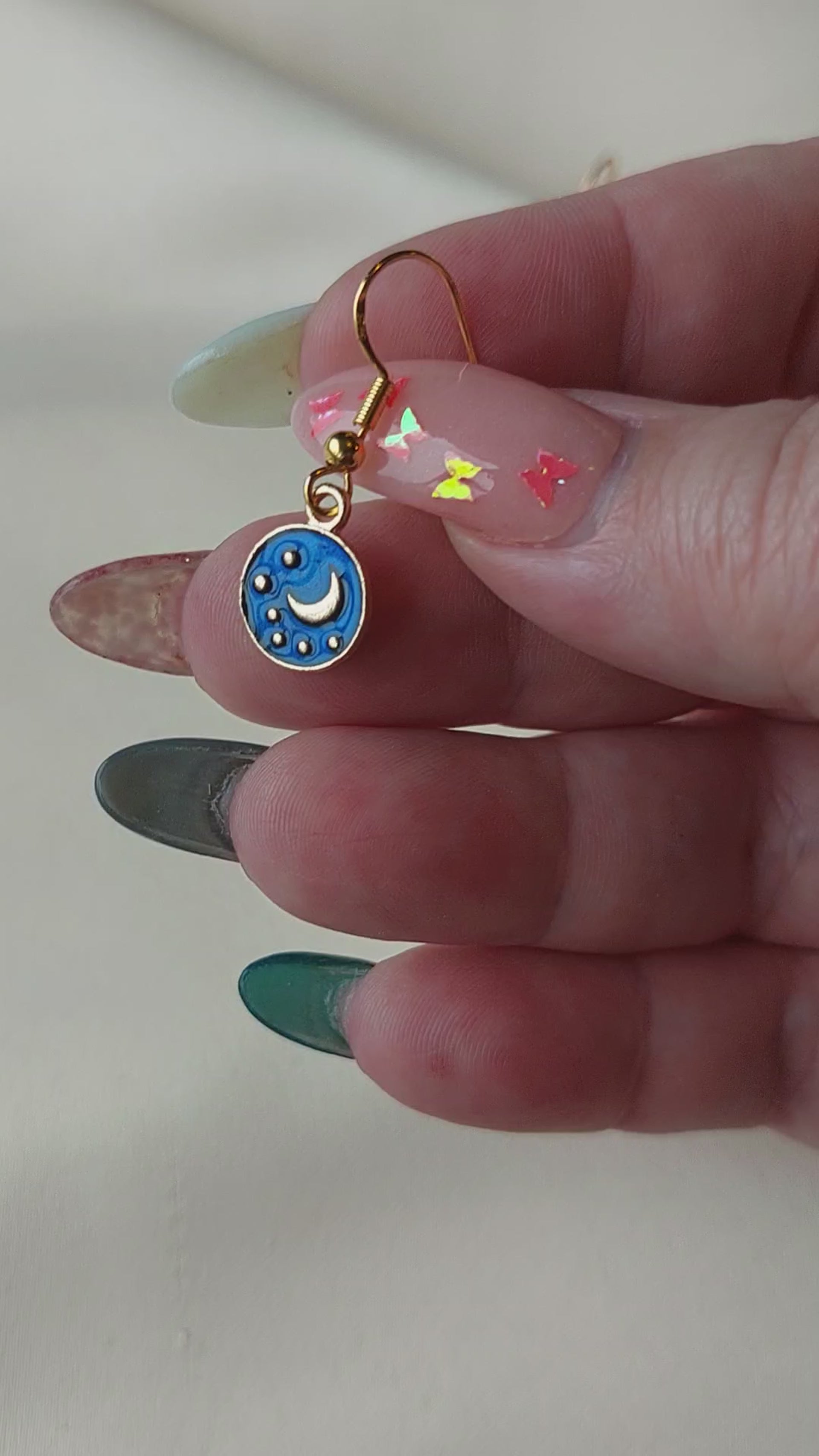Small blue and gold earrings. Circle earring with moon and stars inside. Gold hooks. Earrings laid out on fabric. Hand holding for size comparison. 